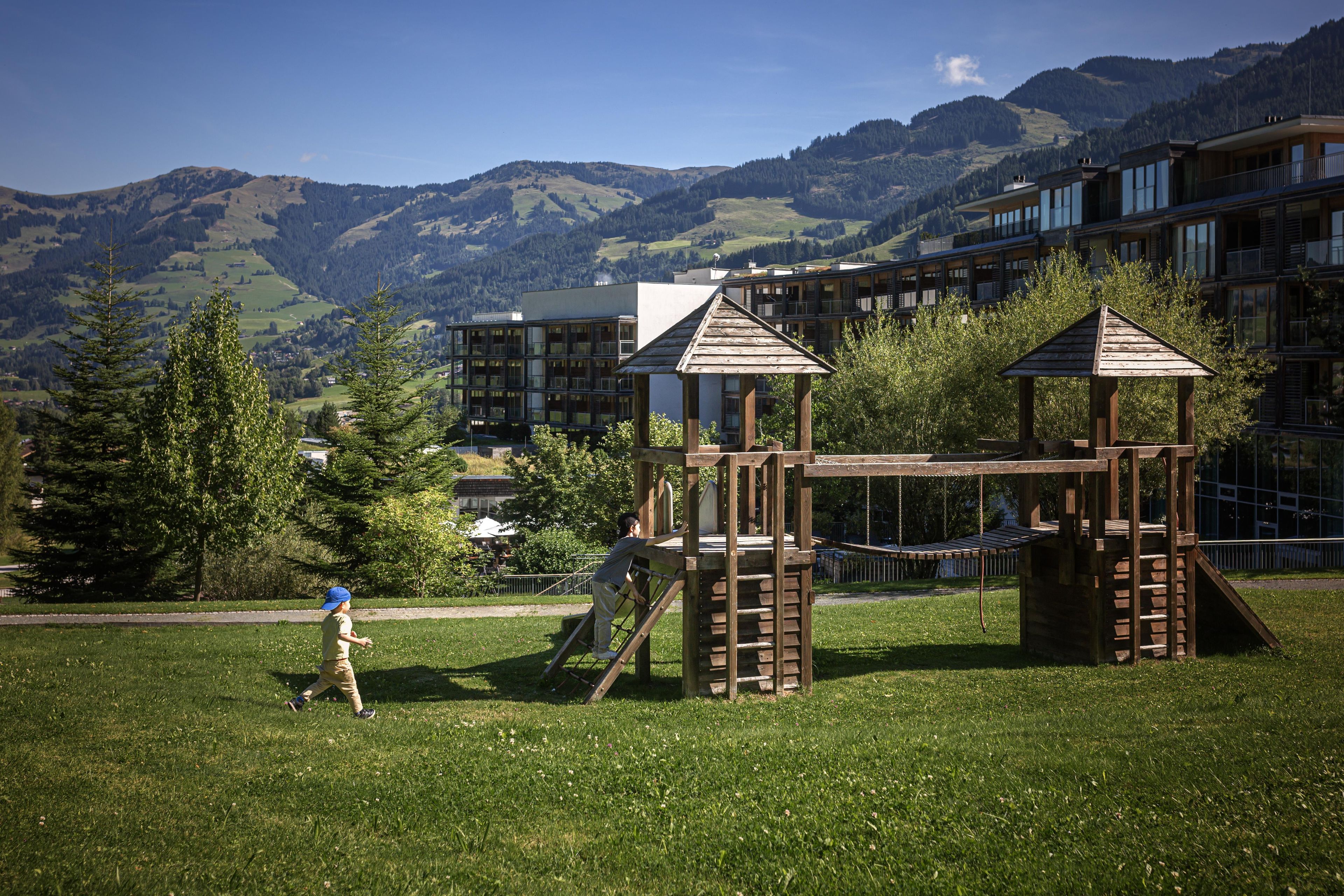 A playground with wooden structures and a child playing, set against a backdrop of a mountainous landscape and modern buildings.
