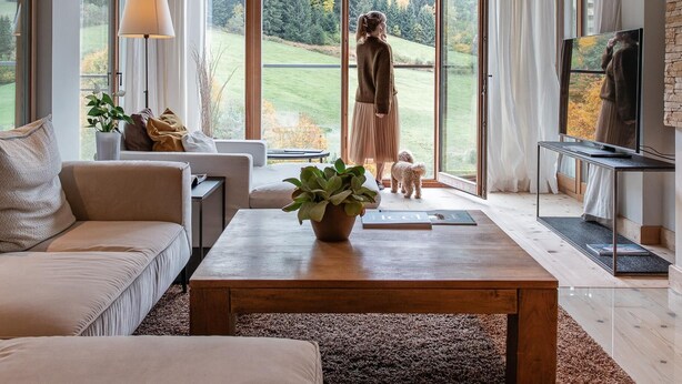A spacious living room with a large window, a wooden ceiling, a sofa, a coffee table, and a television. - Kempinski Hotel Das Tirol (Jochberg)