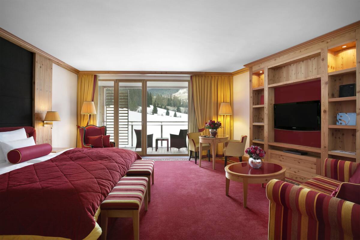 A hotel room with a large bed, a sofa, a TV, a small table, and a balcony with a view of snow-covered mountains.