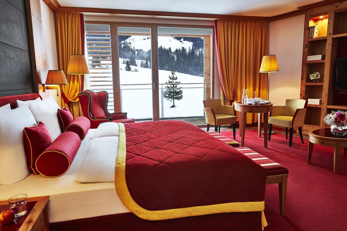 Habitación de hotel con una cama grande, zona de comedor y vistas a un paisaje nevado a través de las ventanas.