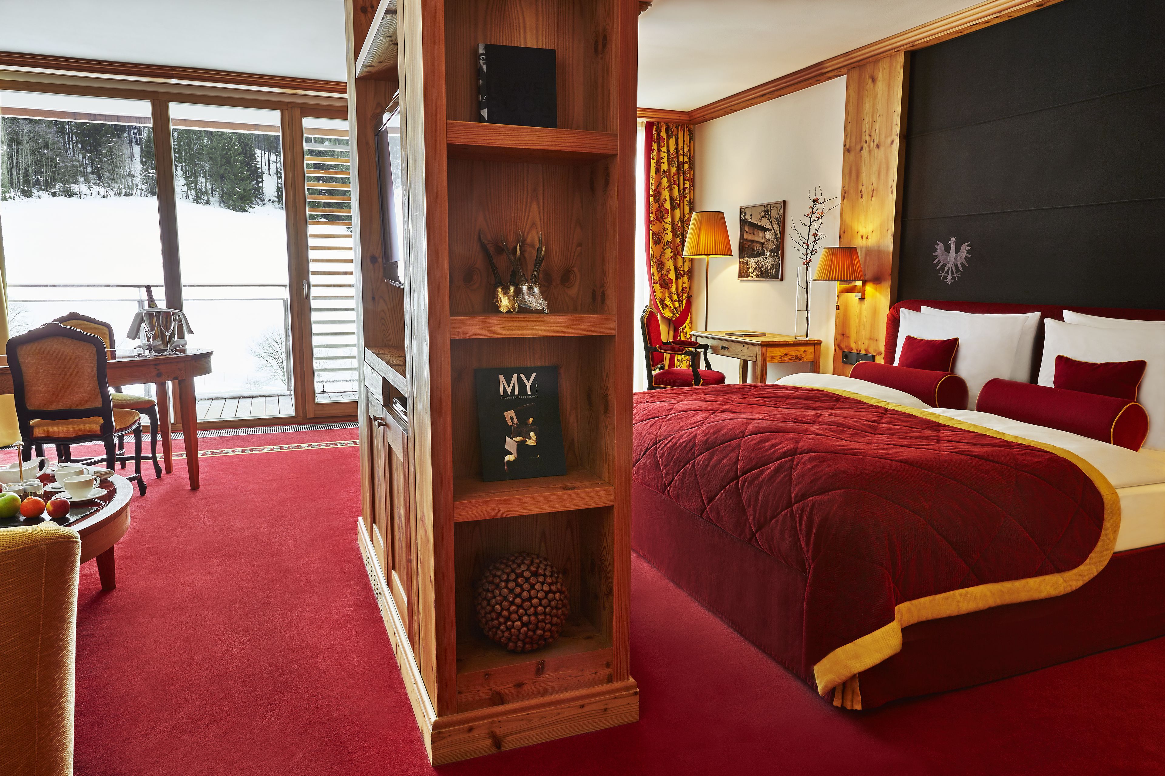 Ein Zimmer mit einem großen Bett, einem Holzbücherregal, einer Essecke mit Tisch und Stühlen und Blick auf eine verschneite Landschaft durch große Fenster.