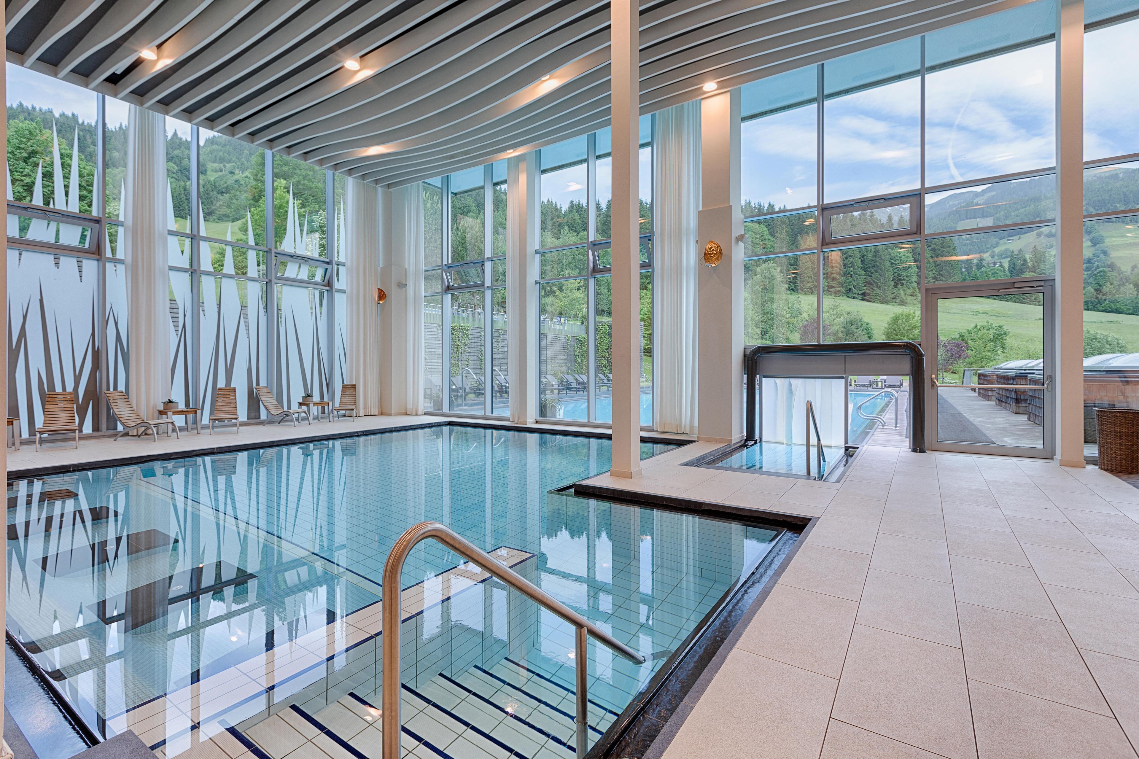 A modern indoor swimming pool with clear glass walls, wooden deck chairs, and a view of a mountainous landscape.