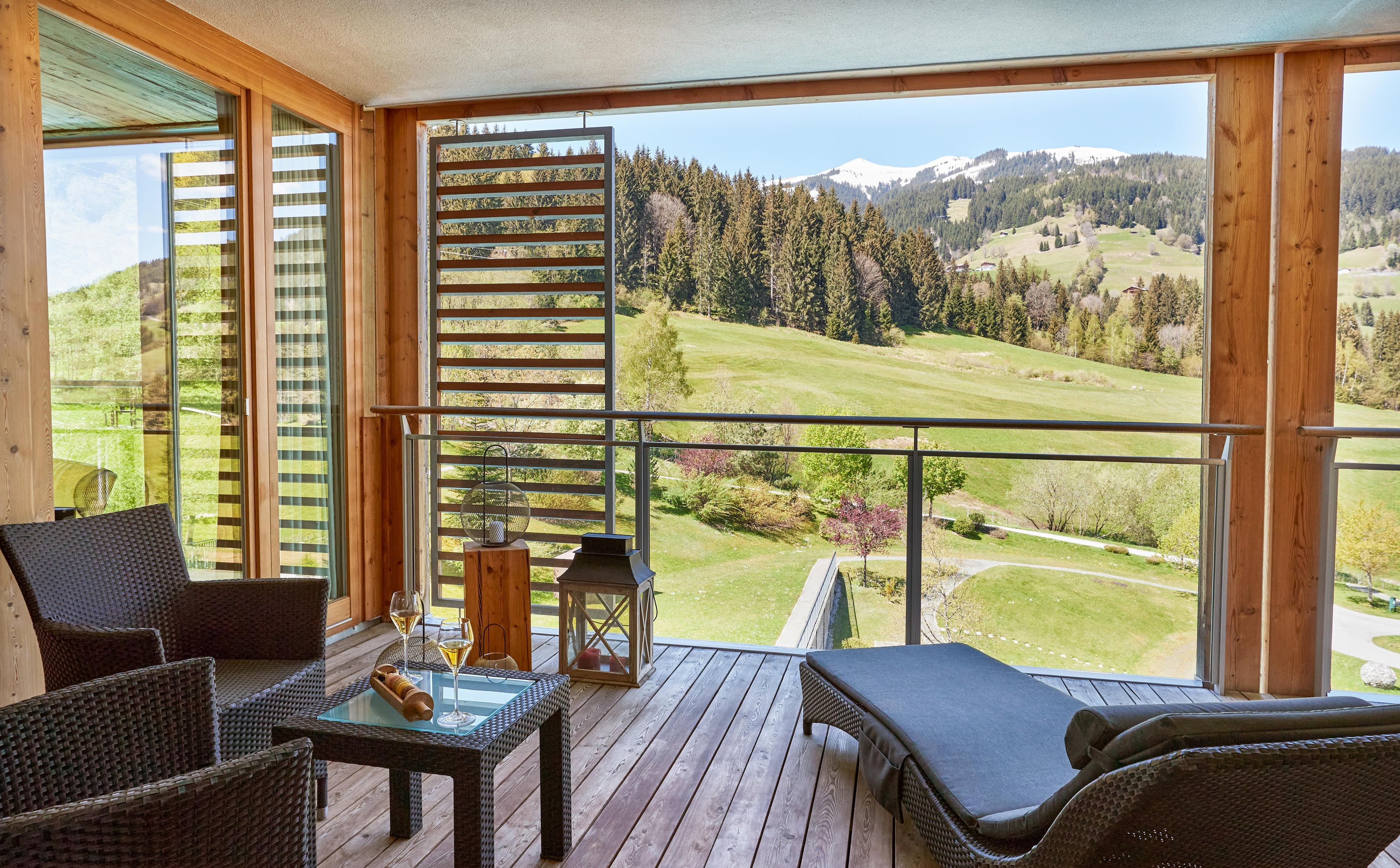 Ein Balkon mit Korbmöbeln, einem Glastisch und Blick auf eine grüne Landschaft mit Bäumen und einem Berg.
