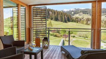 Ein Balkon mit Korbmöbeln, einem Glastisch und Blick auf eine grüne Landschaft mit Bäumen und einem Berg.