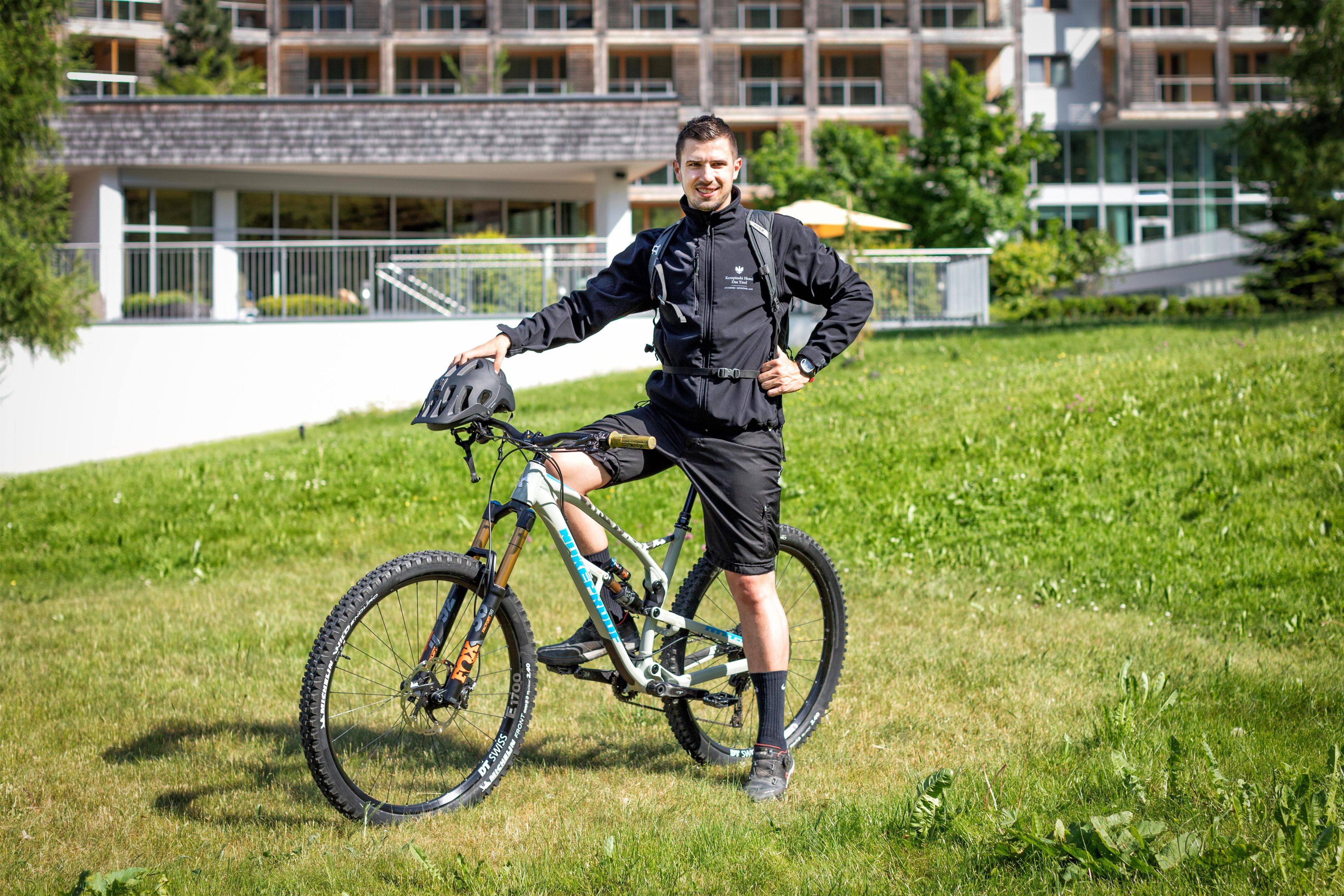 Una persona de pie al lado de una bicicleta de montaña en un campo cubierto de hierba, con un edificio moderno al fondo.