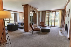 A spacious living room with a large window, a fireplace, and a wooden entertainment center. - Kempinski Hotel Das Tirol (Jochberg)