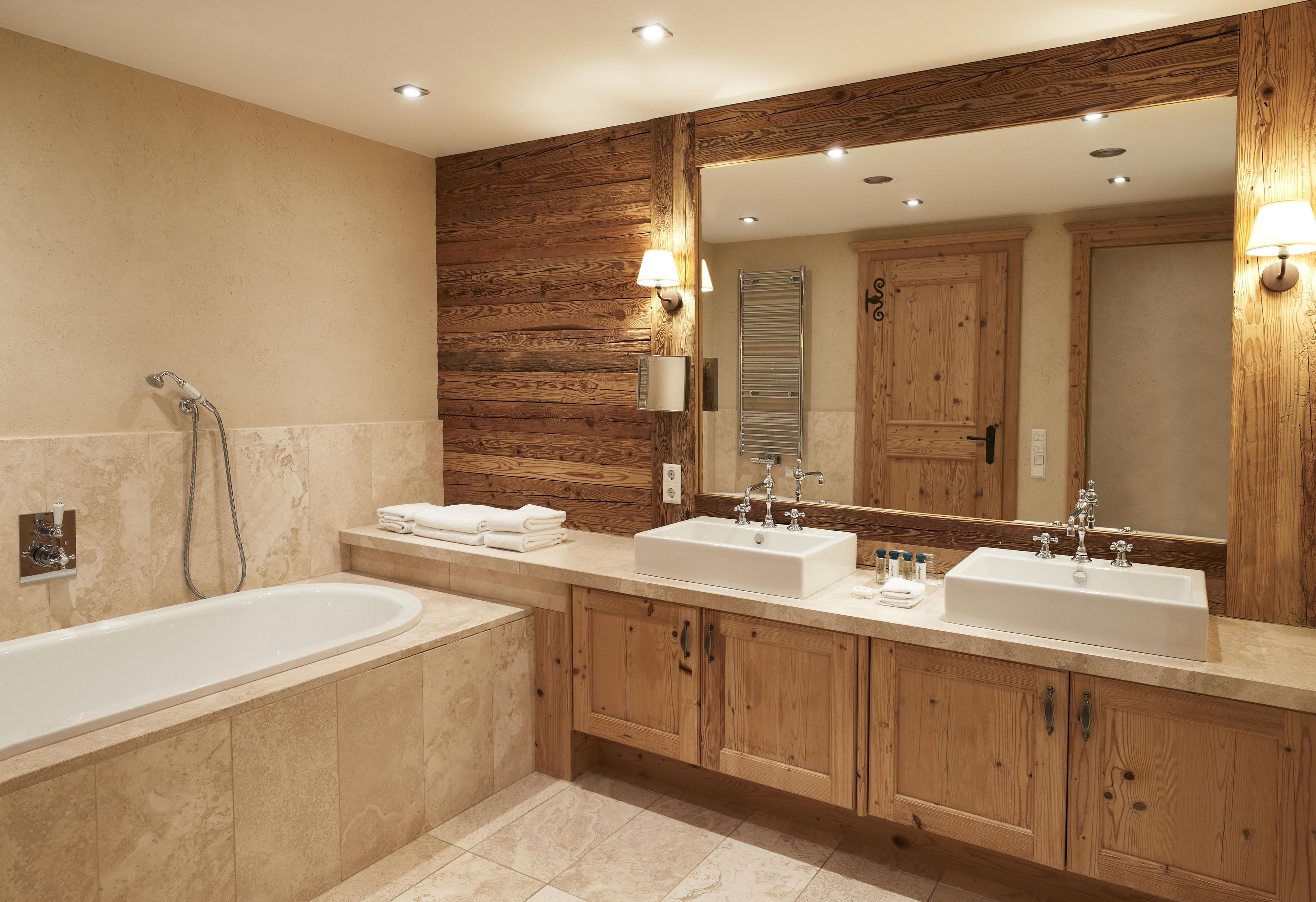 A modern bathroom with a large mirror, two sinks, a bathtub, wooden paneling, and recessed lighting.