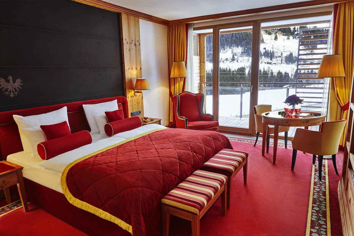 A hotel room with a large bed, a wooden bench, a desk, and a chair. There is a window with a view of a snowy mountain.