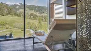 A room with a large window offering a view of a green landscape and mountains. There is a cushioned chair with a towel on it. - Kempinski Hotel Das Tirol (Jochberg)