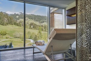 Ein Zimmer mit einem großen Fenster, das einen Blick auf eine grüne Landschaft und Berge freigibt. Ein gepolsteter Stuhl mit einem Handtuch darauf.
