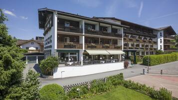 Hotel features a modern design with a flat roof, balconies, and a ground-floor outdoor seating area surrounded by lush greenery.