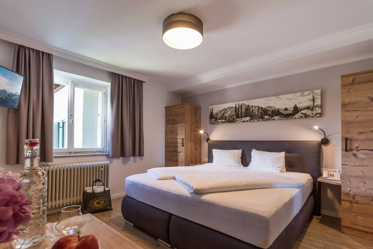 A neatly arranged hotel room with a large bed, a wooden headboard, a painting above the bed, a bedside table with a lamp, and a window with curtains.