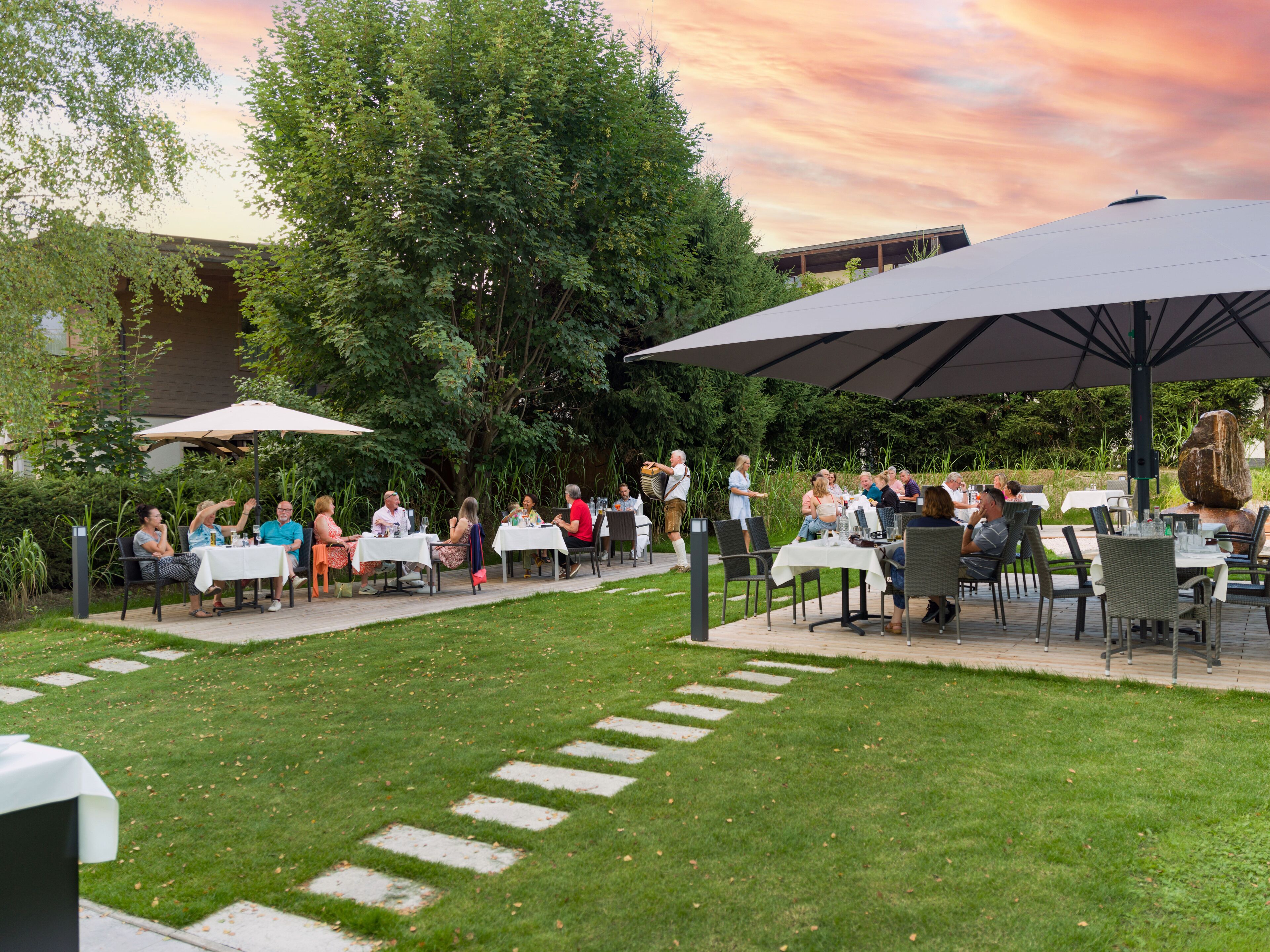 Un espace extérieur aménagé pour les repas, avec des tables et des parasols, entouré de verdure et avec un bâtiment en arrière-plan.