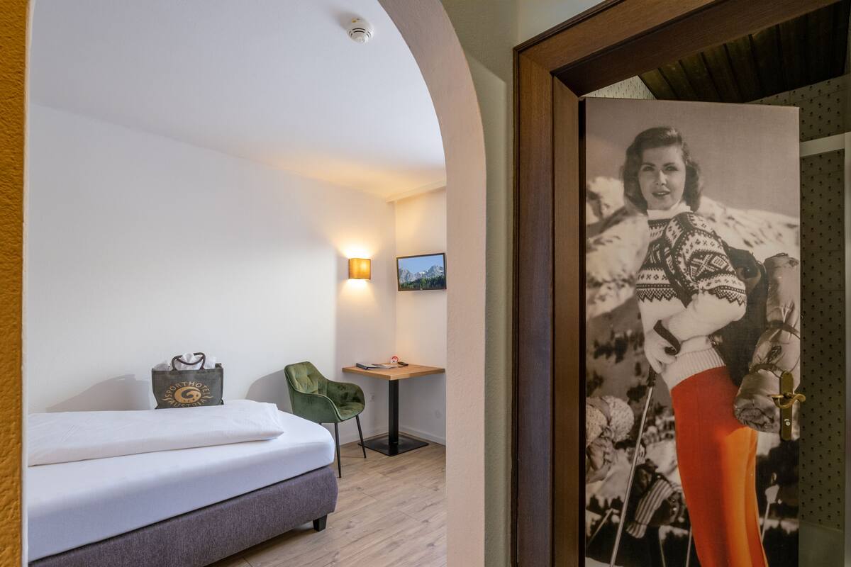 A hotel room with a bed, a green chair, a desk with a lamp, and a framed picture on the wall.