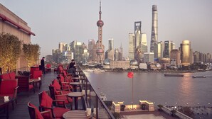 A rooftop terrace with red chairs and tables overlooking a city skyline with prominent buildings and a river. - The Peninsula Shanghai (Shanghai)