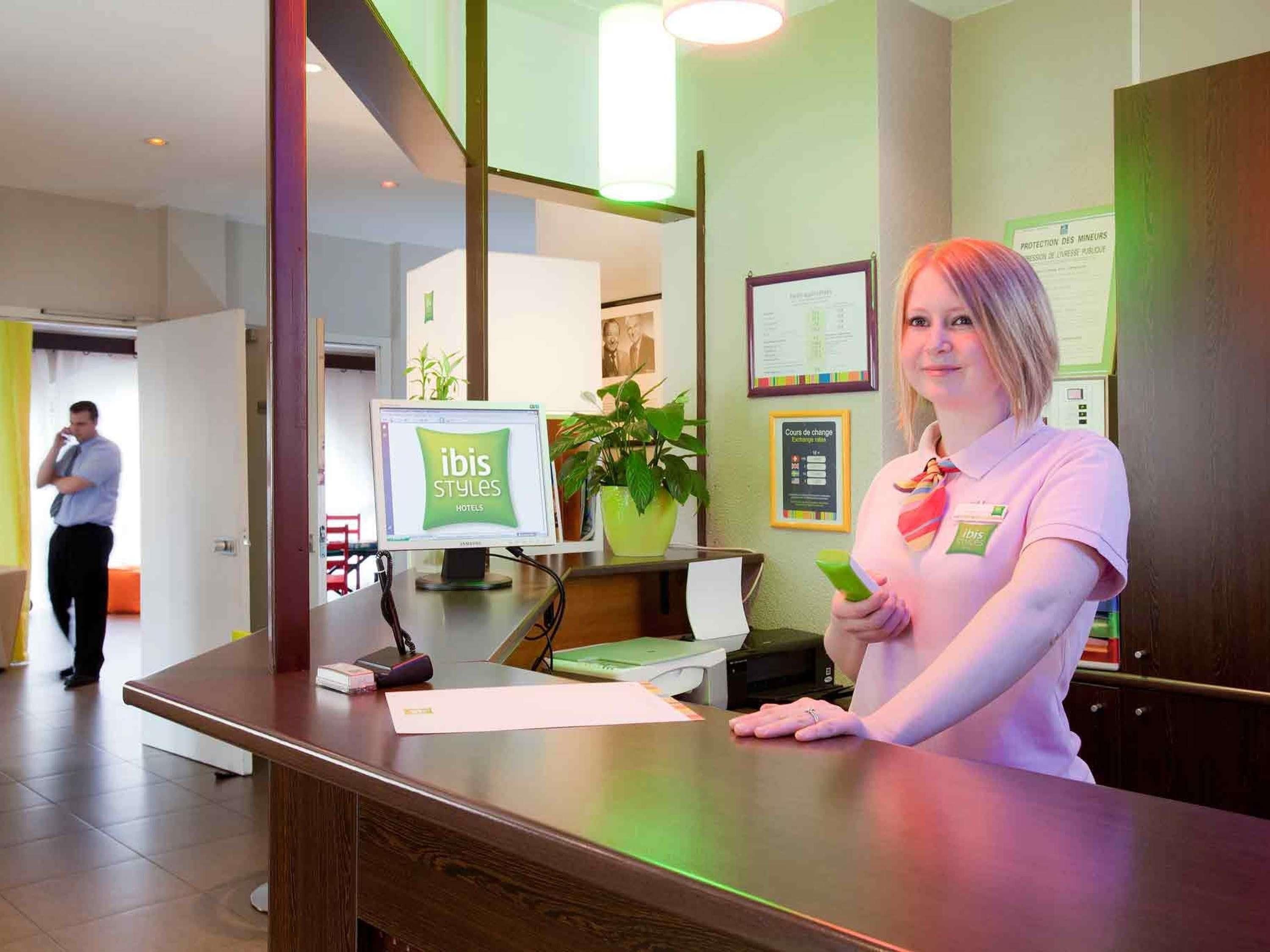 A hotel reception desk with a computer monitor displaying 'ibis Styles Hotels' and a staff member holding a phone.