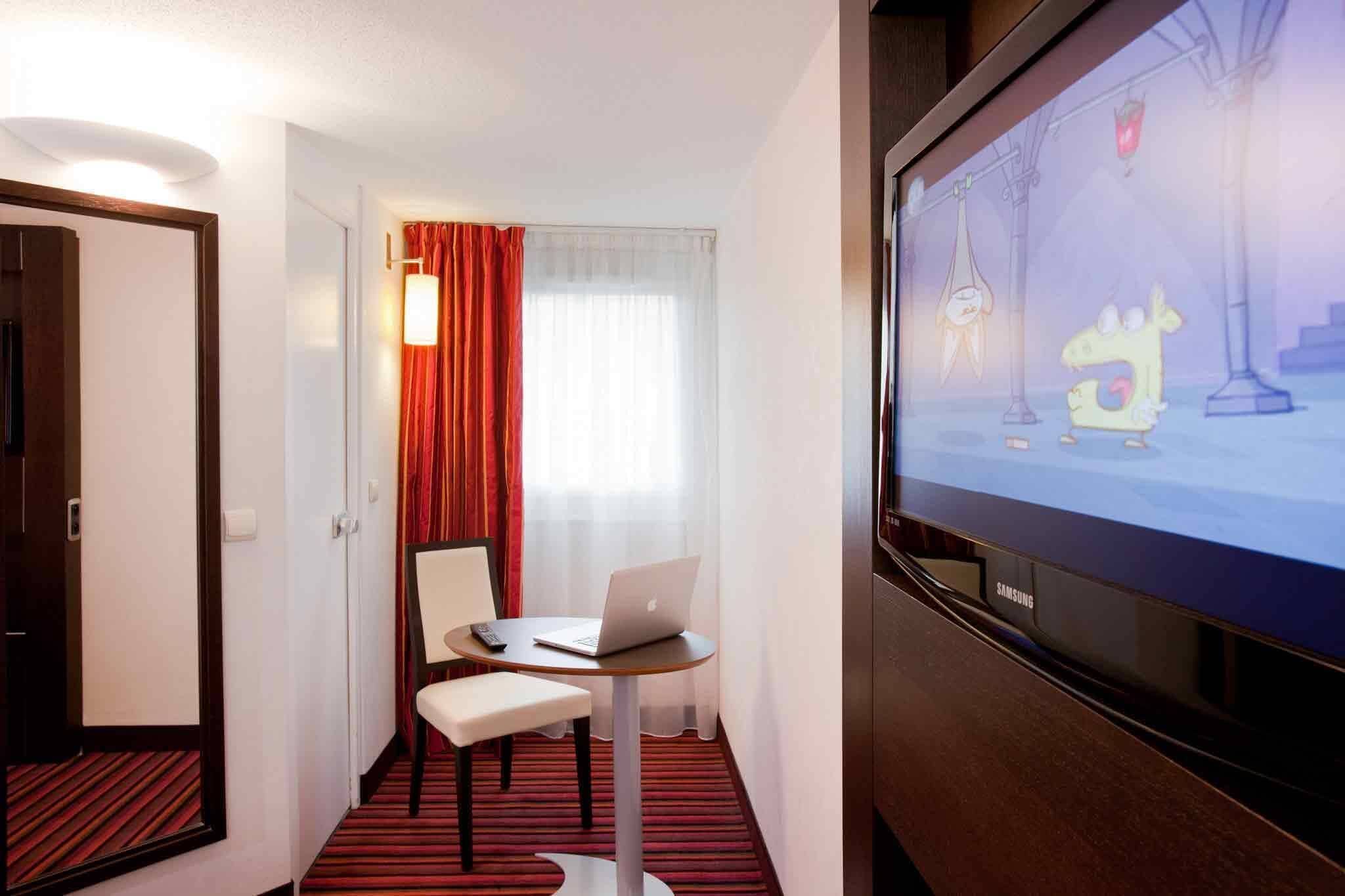 A hotel room with a television, a laptop on a small table, a chair, and a door.