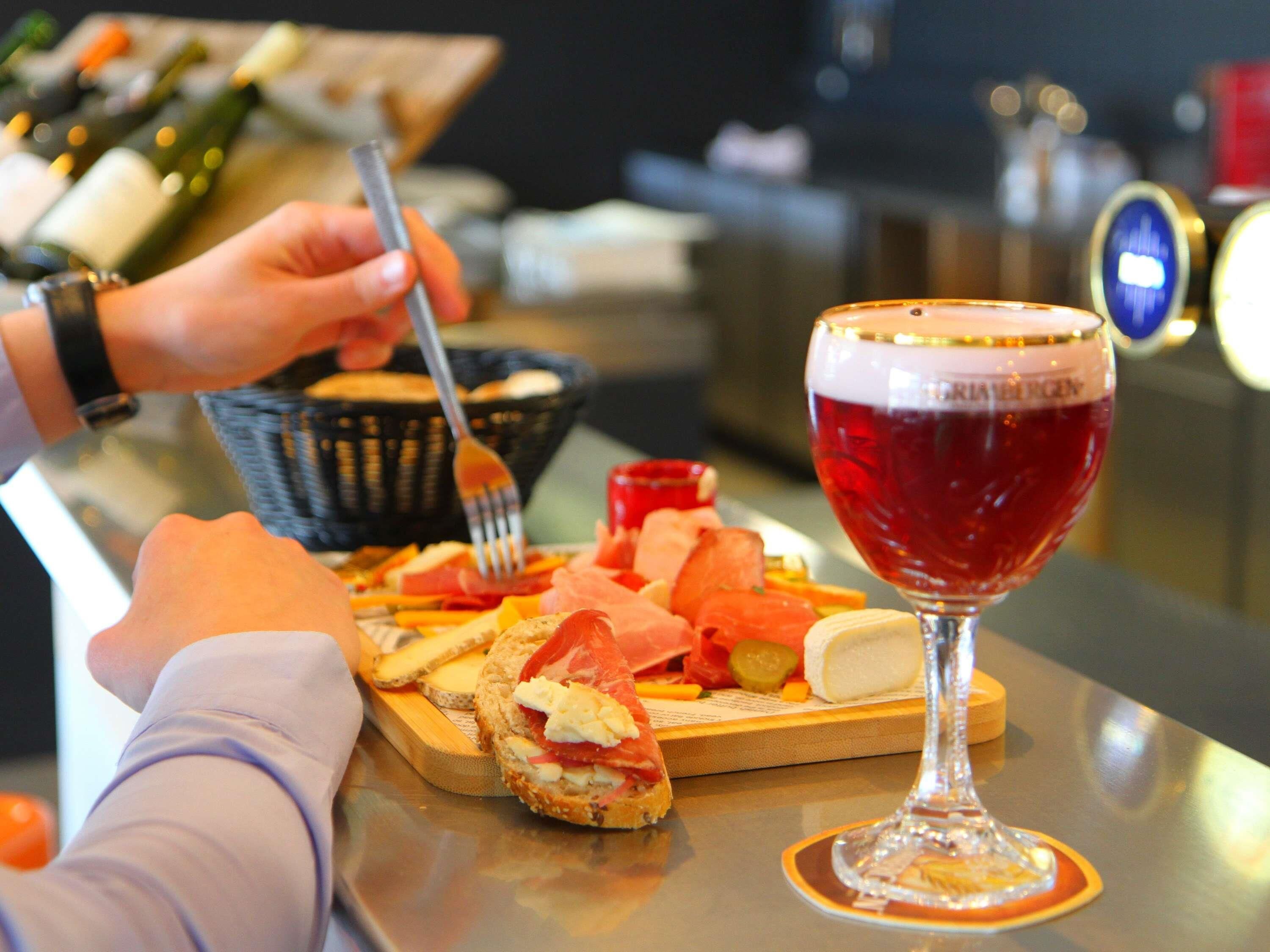 A table with a glass of red wine, a plate of food including bread, cheese, and meat, and a basket of bread rolls.
