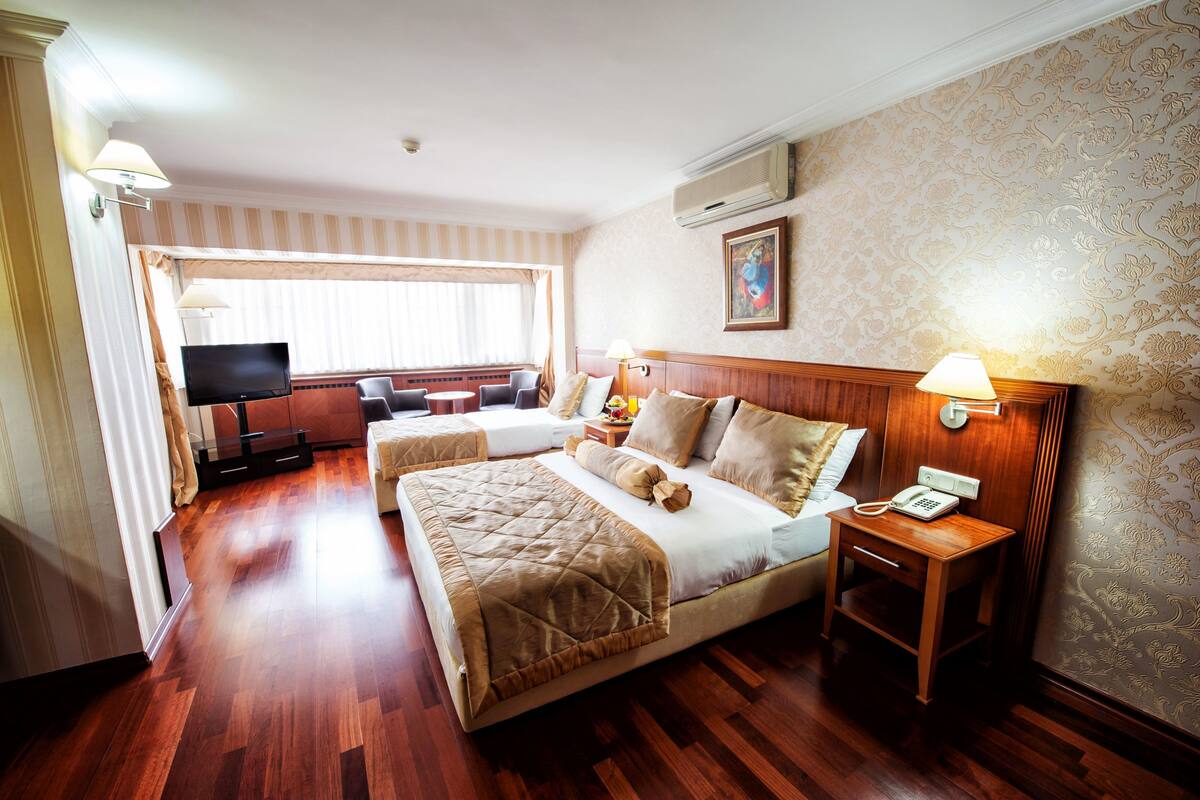 A hotel room with two beds, a TV, a bedside table with a lamp, and a window with curtains.