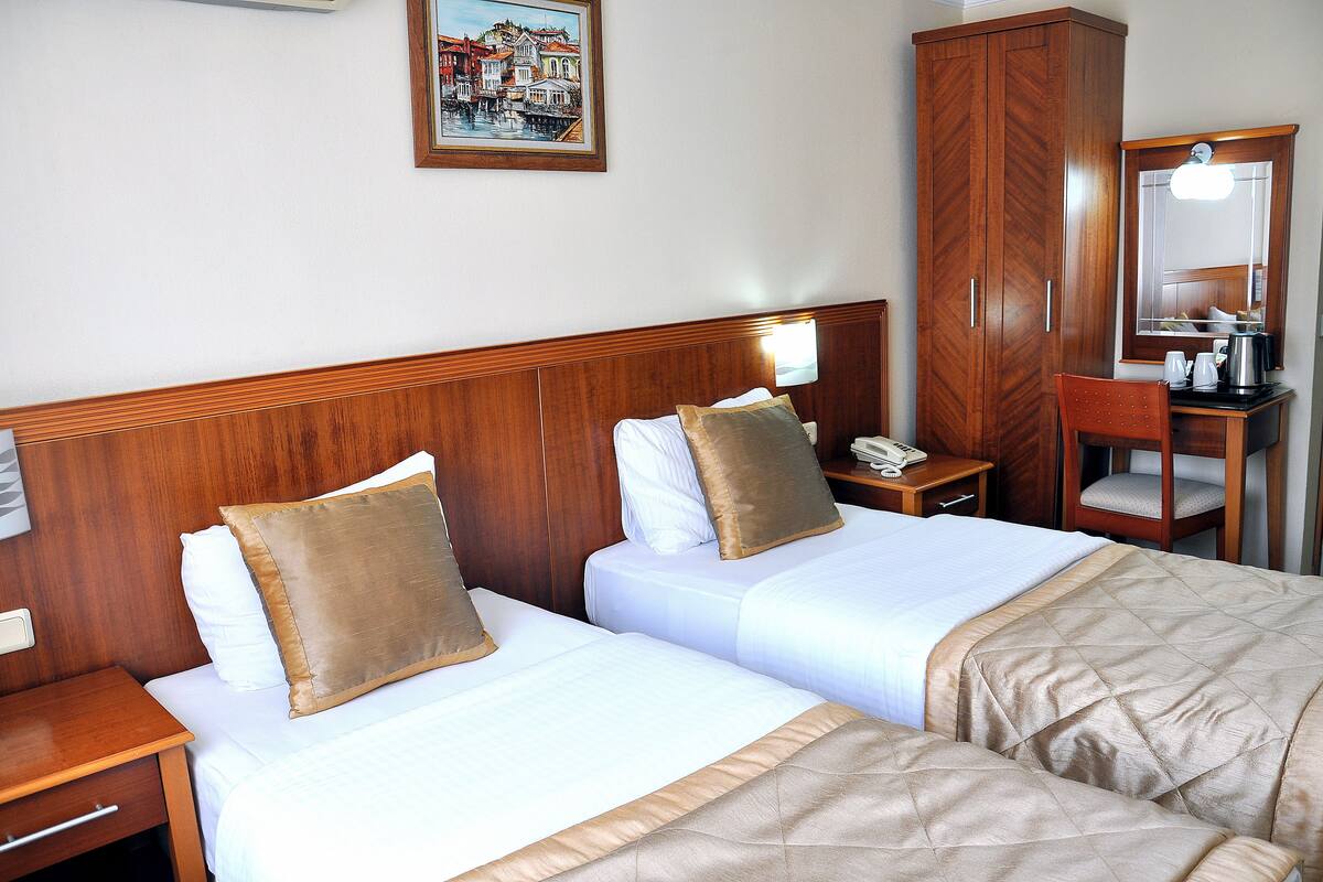 A hotel room with two beds, wooden headboards, a wooden wardrobe, a desk with a telephone, and a mirror.