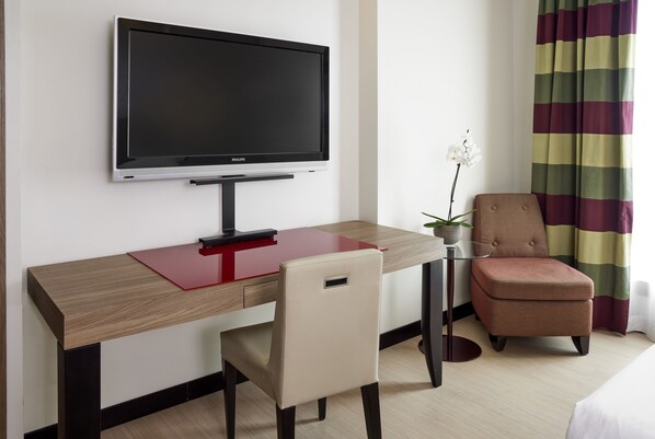 Habitación de hotel con televisor de pantalla plana montado en la pared, un escritorio de madera con superficie roja, una silla color beige, una mesita con una planta y una cortina a rayas.