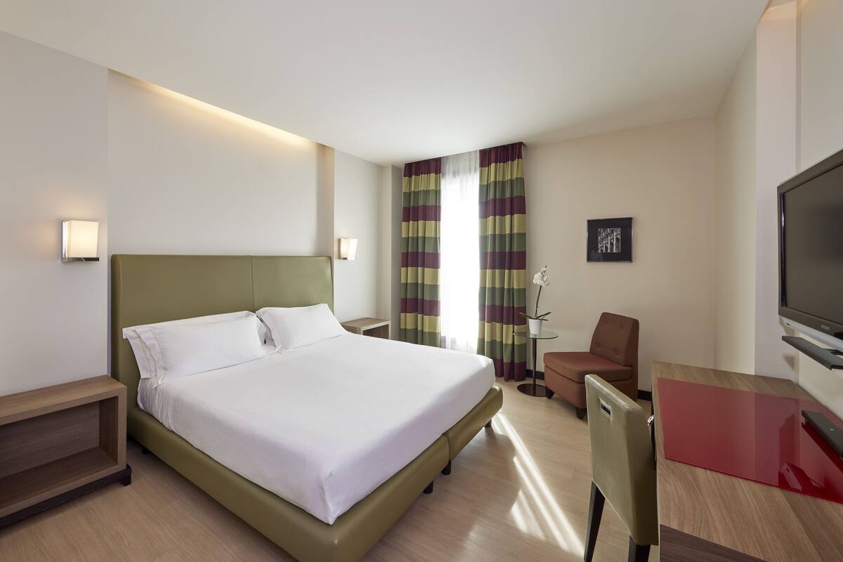Ein Hotelzimmer mit einem großen Bett, einem Schreibtisch mit Fernseher, einem Sessel und einem kleinen Tisch.