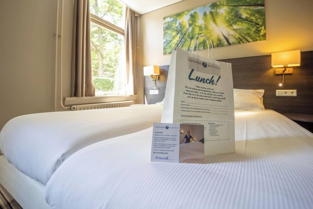 Une chambre d’hôtel avec un lit, une fenêtre avec des rideaux, une lampe fixée au mur et un tableau.
