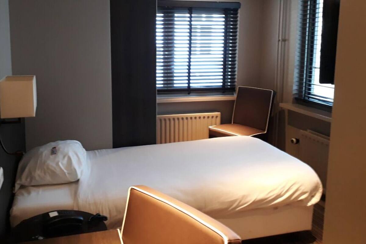 Une chambre d’hôtel avec un lit, une chaise, une table de chevet, une lampe et une fenêtre avec des stores.