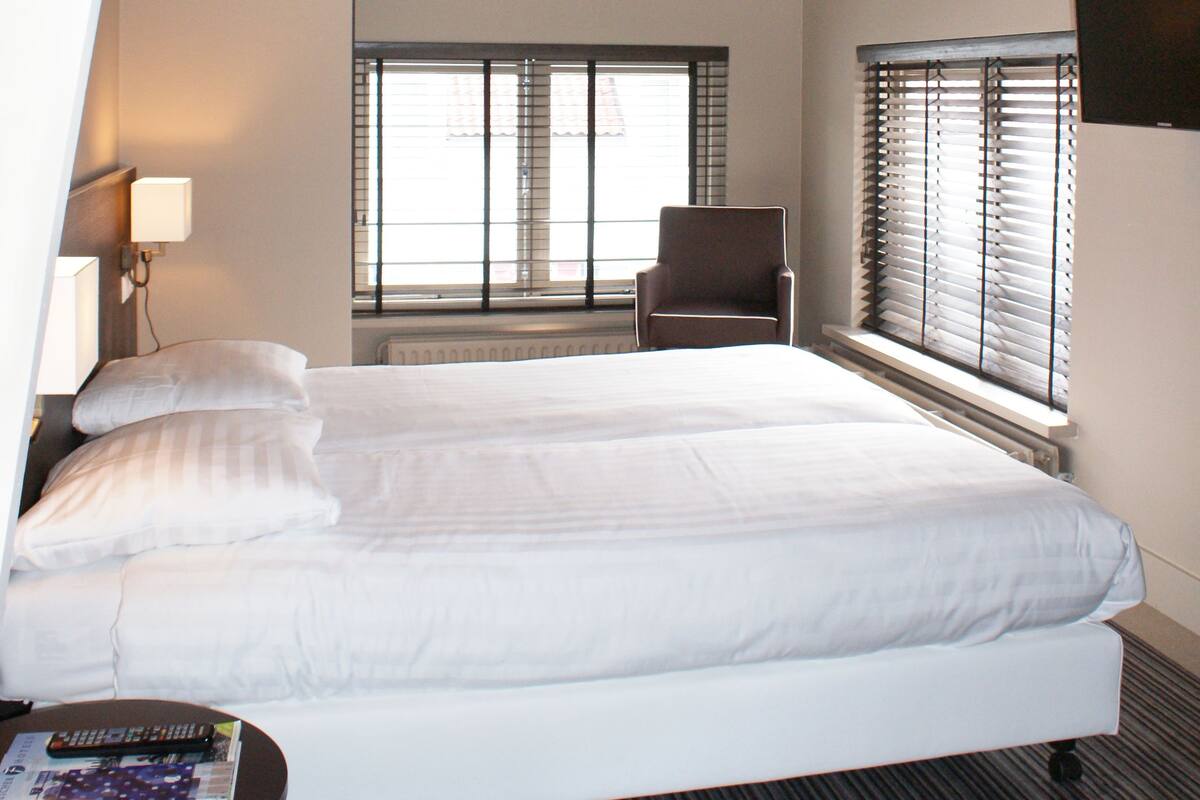 Une chambre d’hôtel moderne avec un grand lit, une chaise et une table. Elle est dotée de fenêtres avec des stores, d’une lampe et d’un luminaire au plafond.