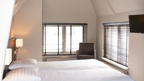 Une chambre d’hôtel moderne avec un grand lit, une chaise et une table. Elle est dotée de fenêtres avec des stores, d’une lampe et d’un luminaire au plafond.