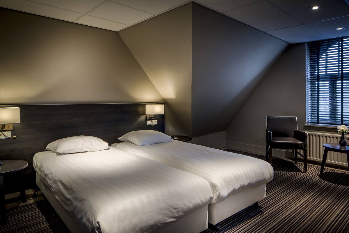 Une chambre d’hôtel avec un grand lit, deux tables de chevet avec des lampes, une chaise et une fenêtre avec des stores.