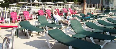 A poolside area with green lounge chairs and pink chairs, some occupied by people.
