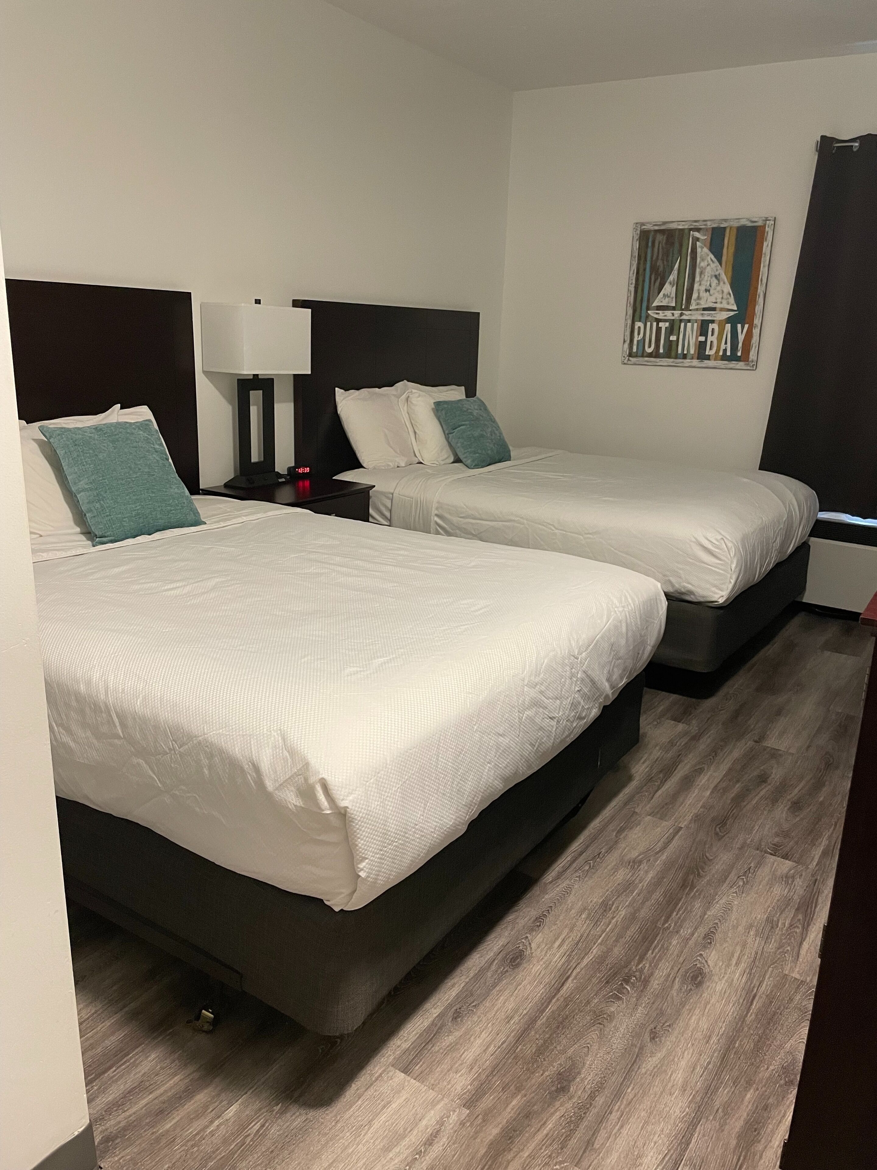 Une chambre d’hôtel avec deux lits, une table de chevet avec une lampe, une horloge murale et un panneau indiquant « Put-in-Bay ».
