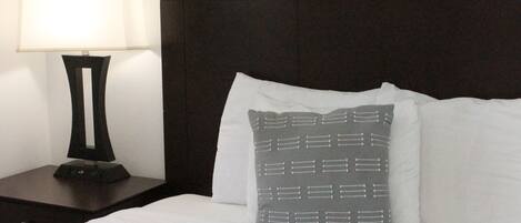 Una cama bien hecha con sábanas blancas, un cabecero oscuro y un cojín estampado en gris. Una lámpara de noche está sobre una mesita junto a la cama.