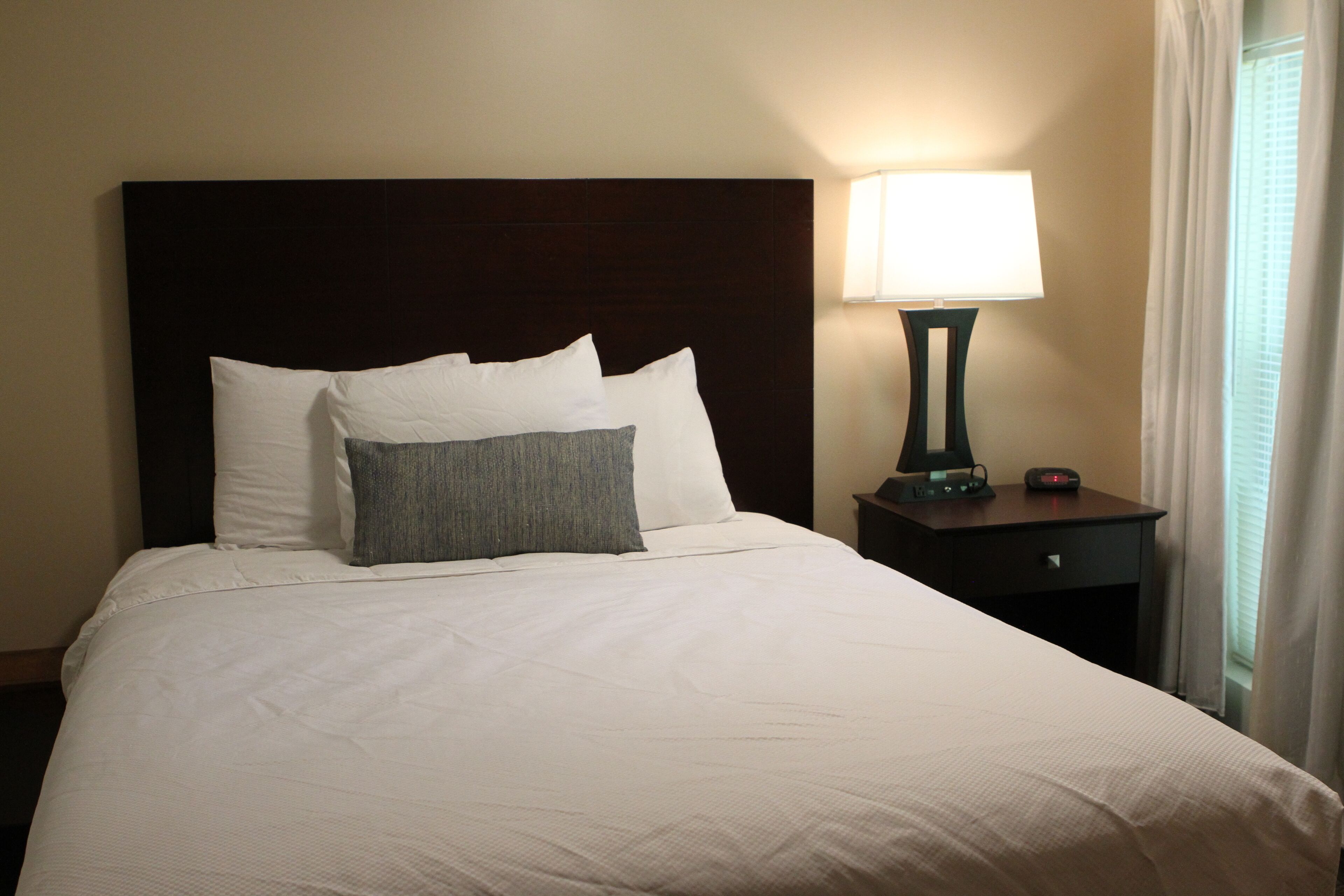 A neatly made bed with white bedding and a dark headboard. There is a bedside table with a lamp and a small clock.