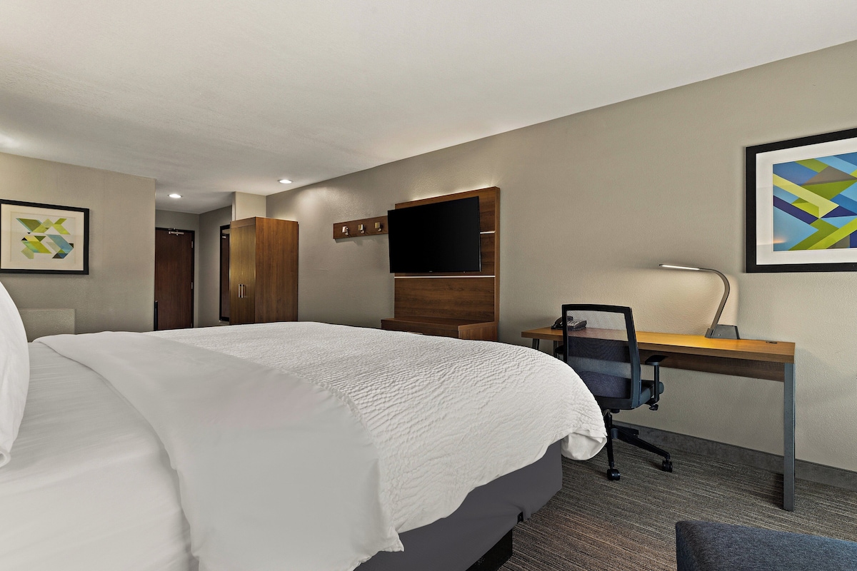 Quarto de hotel com uma cama grande, uma escrivaninha com cadeira, uma TV de tela plana e um quadro abstrato emoldurado.