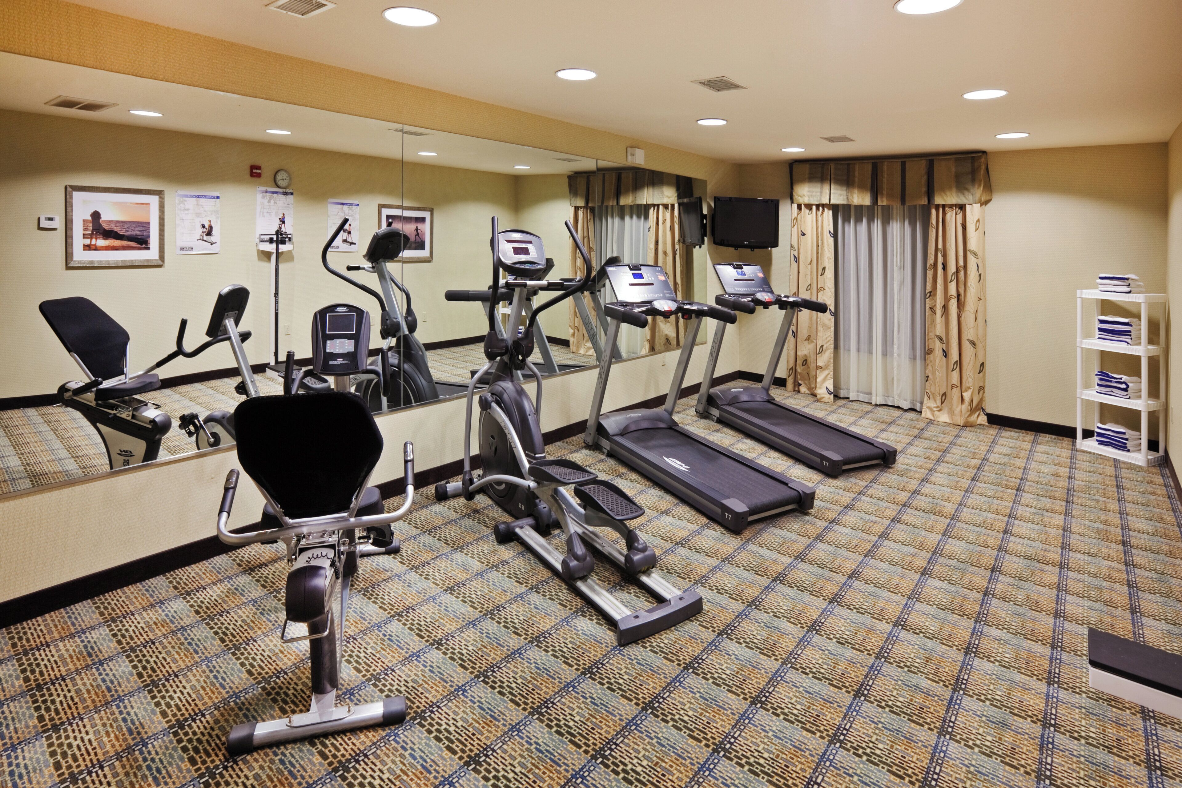 Gimnasio de hotel con equipo de ejercicio, un espejo y suelo alfombrado.