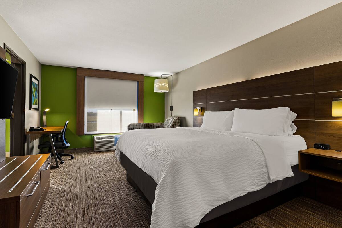 A hotel room with a large bed, a desk, a chair, a window with blinds, and a wall-mounted lamp.