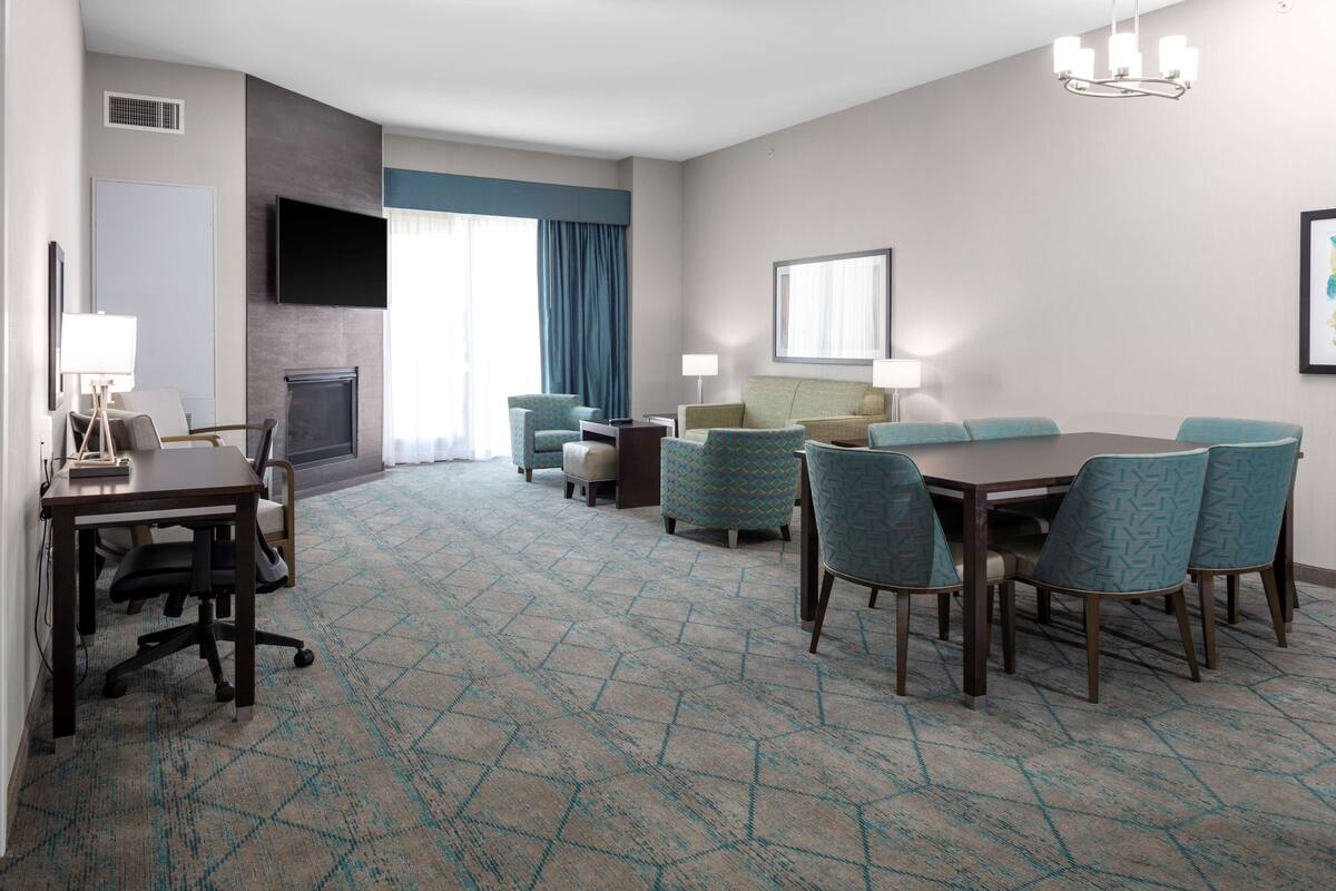 Habitación de hotel con una cama grande, un escritorio con silla, un televisor montado en la pared y una zona de comedor con sillas y mesa.