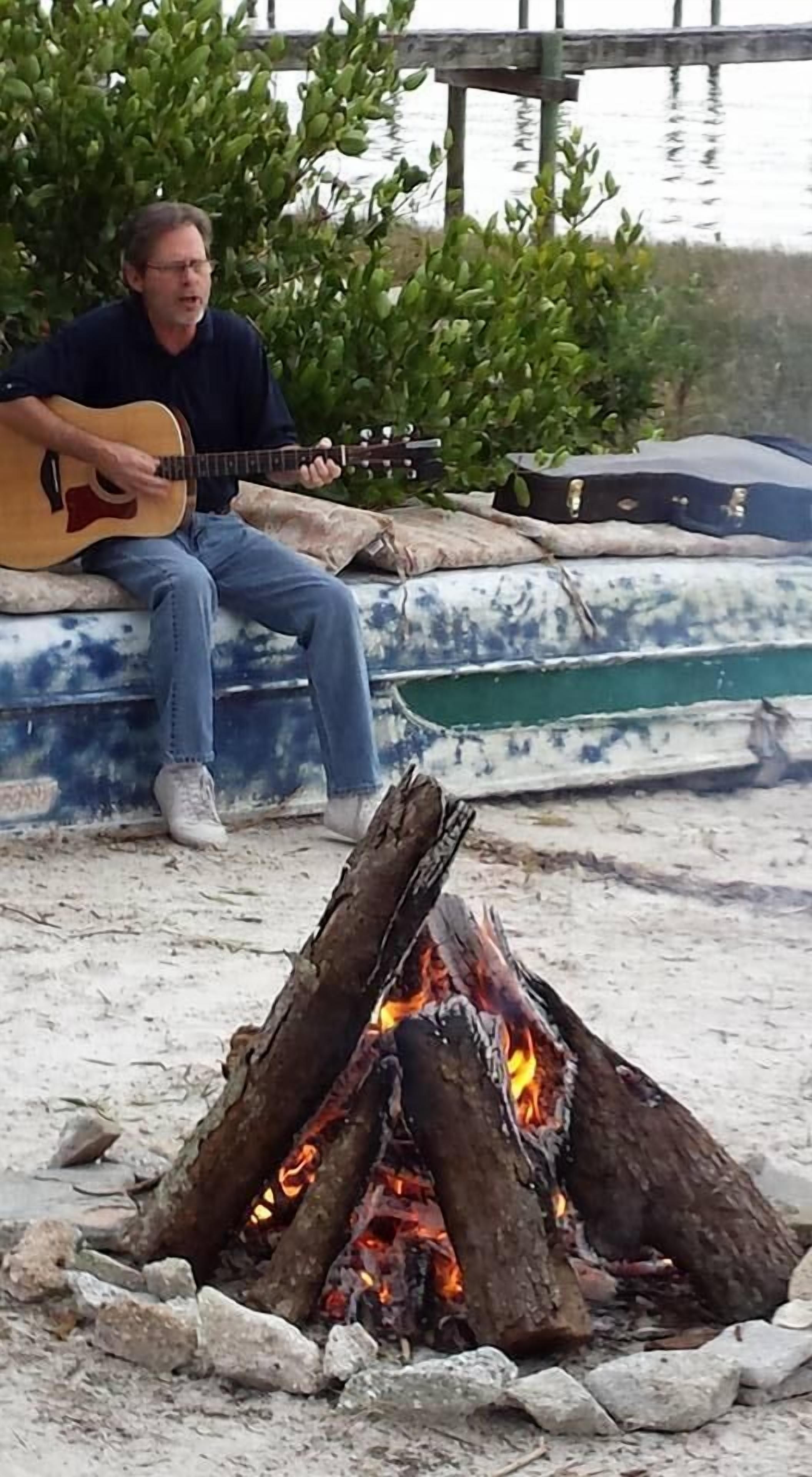 Eine Person spielt akustische Gitarre neben einem Lagerfeuer in der Nähe eines Gewässers.