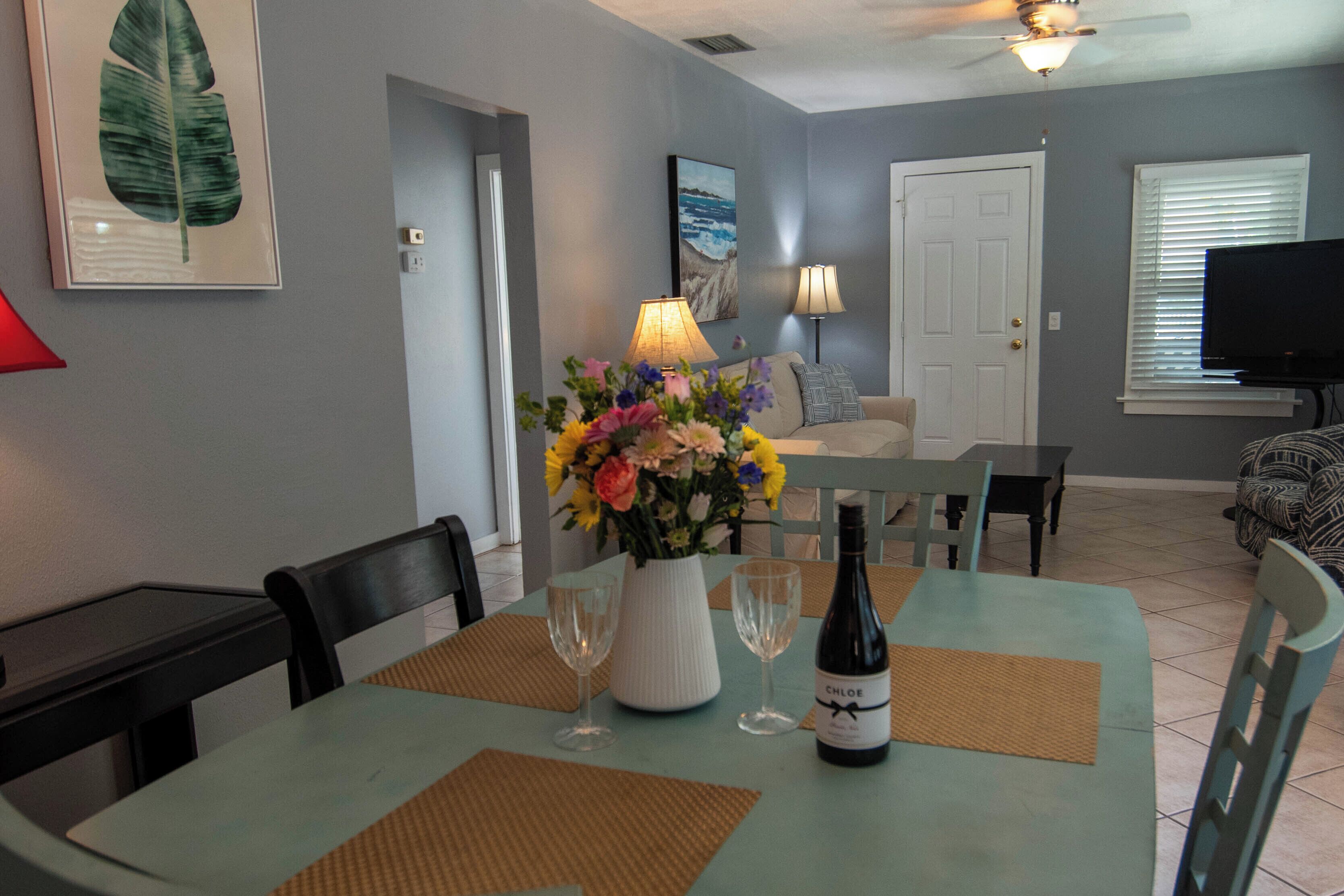 Un coin repas avec une table dressée pour deux personnes, un vase de fleurs et une bouteille de vin.