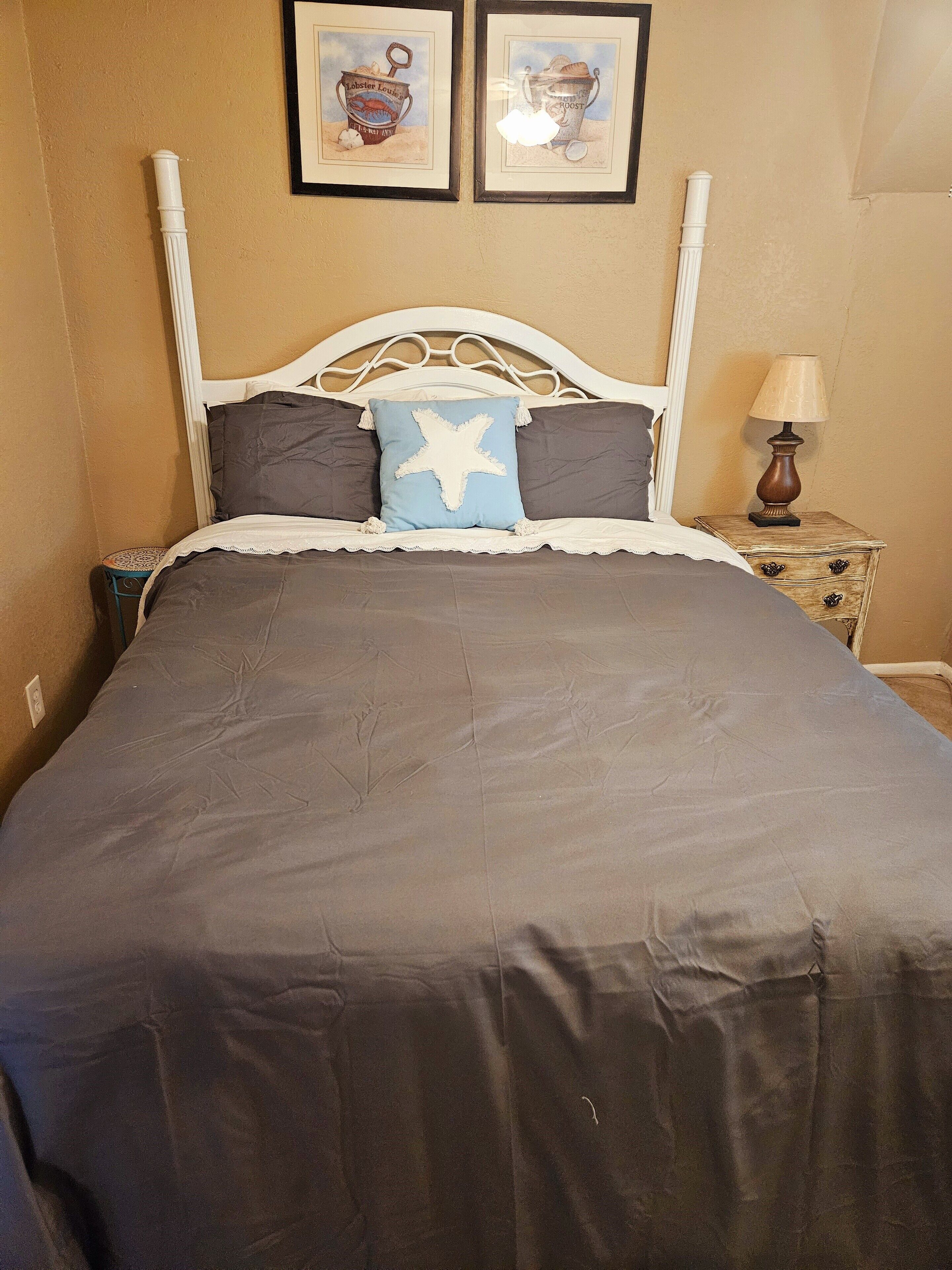 Ein Bett mit weißem Metallgestell, dunkler Bettwäsche und einem sternförmigen Kopfkissen. Über dem Bett hängen zwei gerahmte Bilder an der Wand.