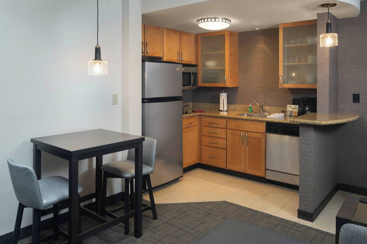A modern kitchen with wooden cabinets, stainless steel appliances, a granite countertop, and a dining area with two chairs.