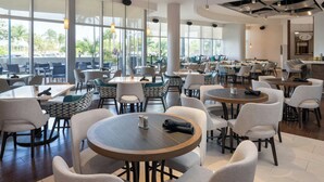 Un restaurant moderne avec des tables et des chaises rondes, de grandes fenêtres et une vue sur les palmiers.