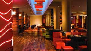 Un hall d’hôtel moderne avec un plancher en bois, des sièges rouges et verts, et un plafond doté de panneaux lumineux.