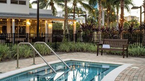 Une piscine dont la profondeur maximale est indiquée à 3 pieds, entourée d’une terrasse en briques, de palmiers et d’un banc.