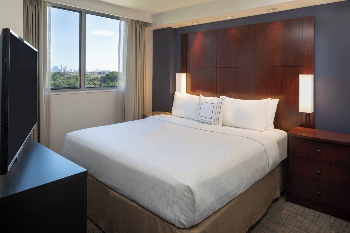 A hotel room with a large bed, a TV on a stand, a nightstand, and a window with a city view.