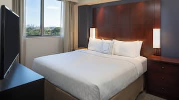Habitación de hotel con una cama grande, un televisor sobre un soporte, una mesita de noche y una ventana con vistas a la ciudad.