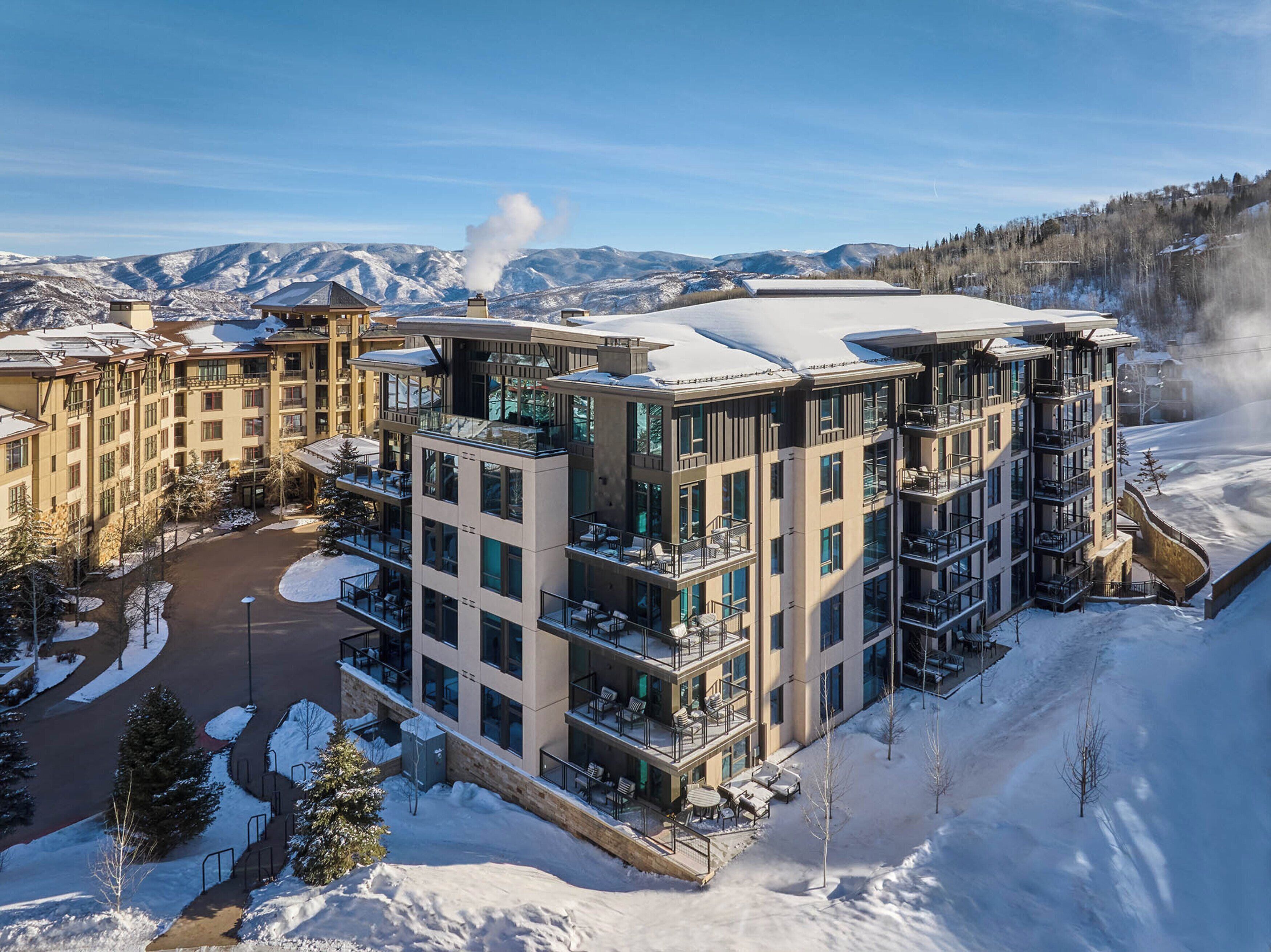 A multi-story hotel with balconies, a snow-covered roof, and a view of mountains.