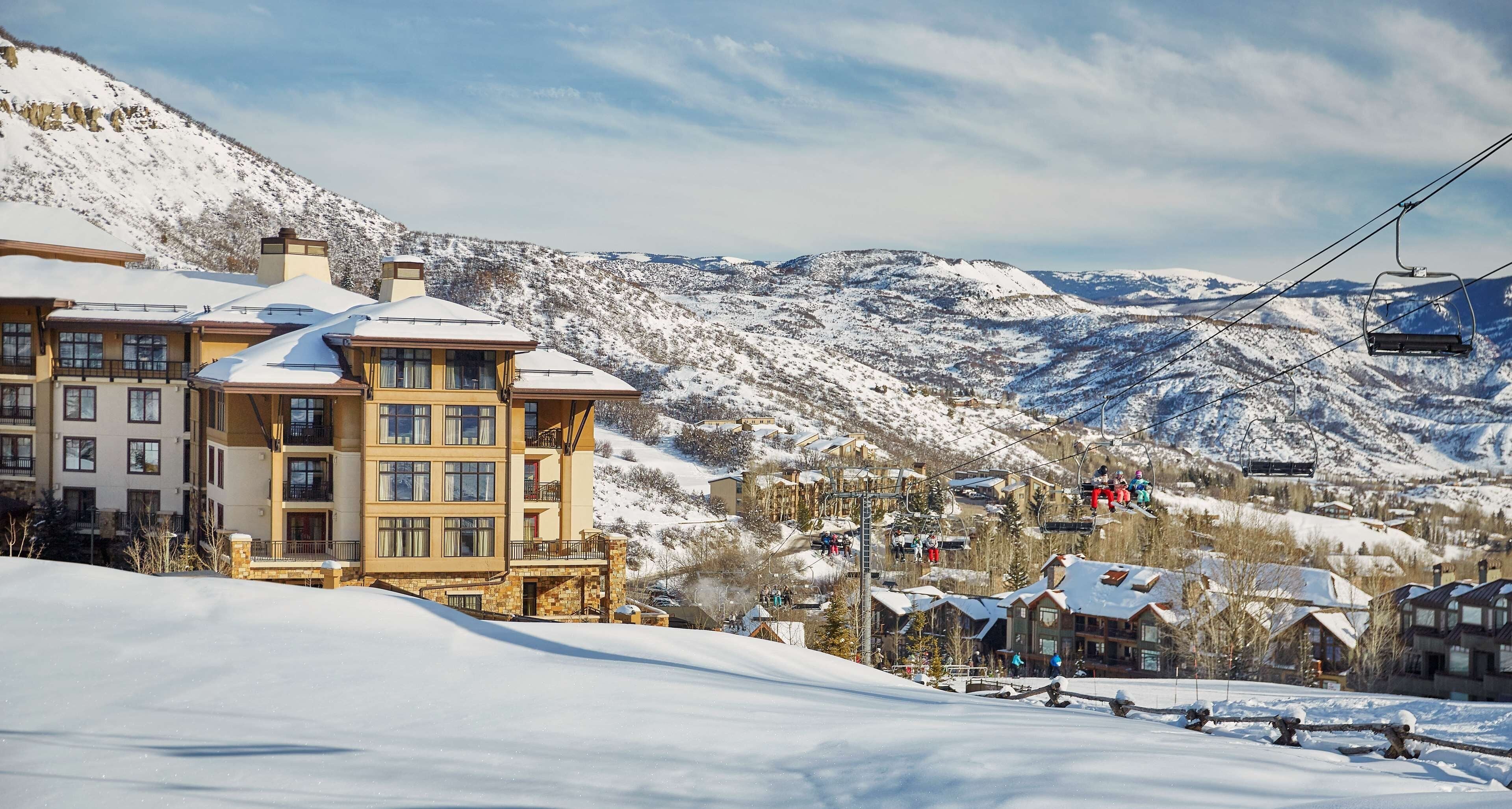 A snow-covered mountain resort with multi-story buildings, ski lifts, and a clear view of the surrounding mountains.